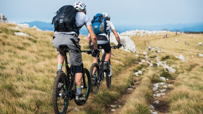 Two riders on e-bikes navigating a rocky trail through open grassy terrain