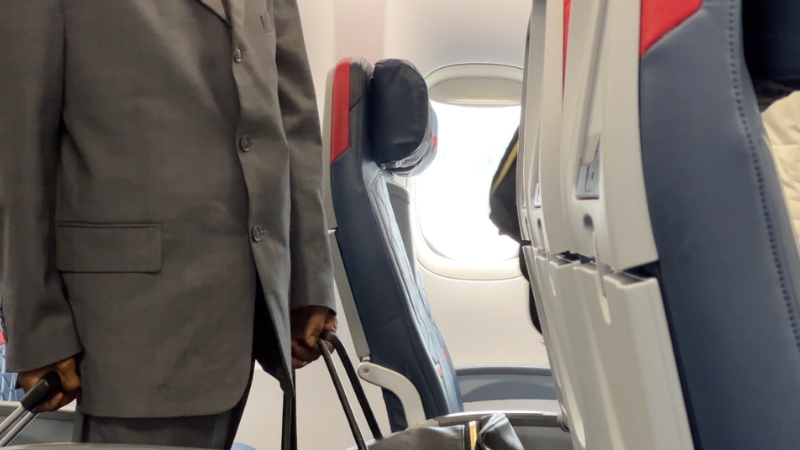 A passenger in a suit stands in an airplane aisle with a carry-on bag, preparing for budget-friendly travel