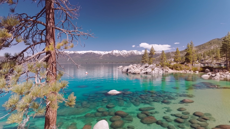 Clear turquoise water and smooth rocks fill Lake Tahoe with snowy mountains rising in the background