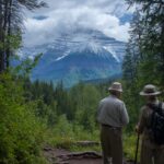Two elderly hikers with walking sticks stand on a forested trail overlooking a snow-dusted mountain under partly cloudy skies