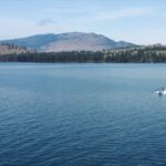 A speedboat moves across the clear blue waters of Flathead Lake with forested hills and mountains in the background