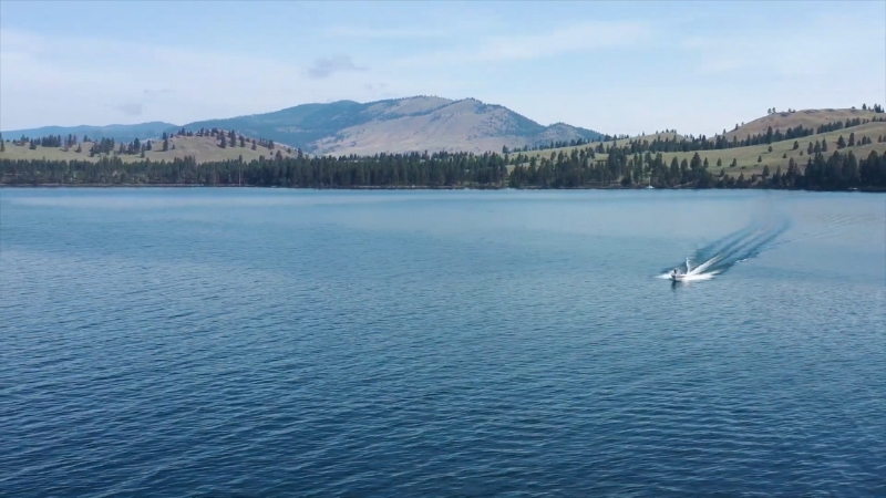 A speedboat moves across the clear blue waters of Flathead Lake with forested hills and mountains in the background
