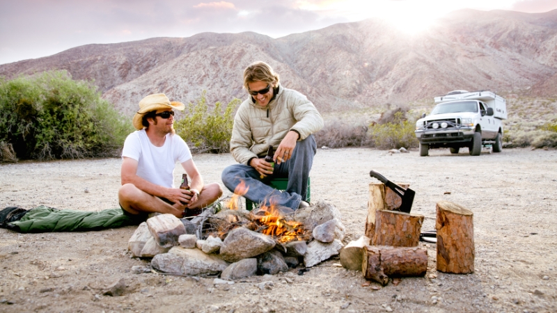 Two men sit by a campfire in a desert area with mountains in the background and a parked off-road vehicle nearby