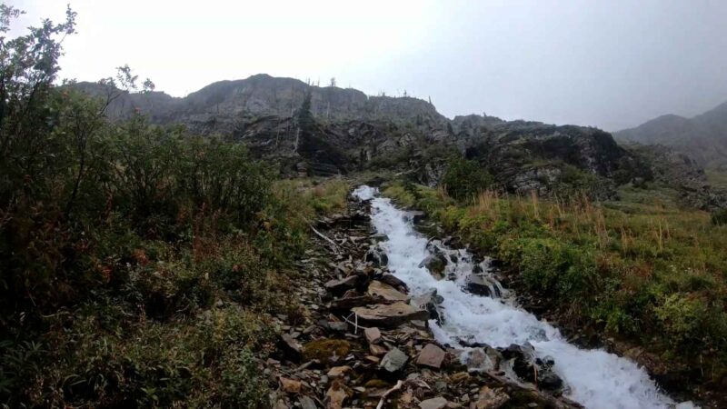 A cascading stream running through rocky terrain and dense green vegetation with steep mountains in the background under a cloudy sky