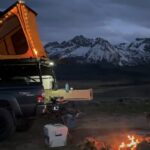A camper sits by a campfire next to a truck with a rooftop tent
