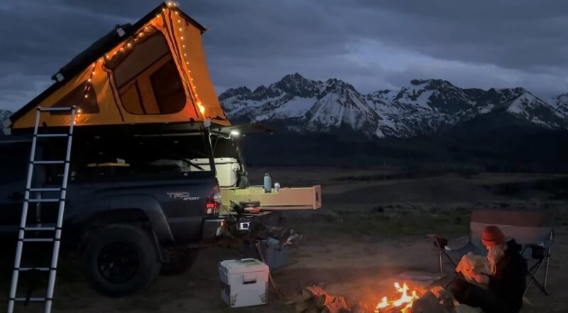 A camper sits by a campfire next to a truck with a rooftop tent