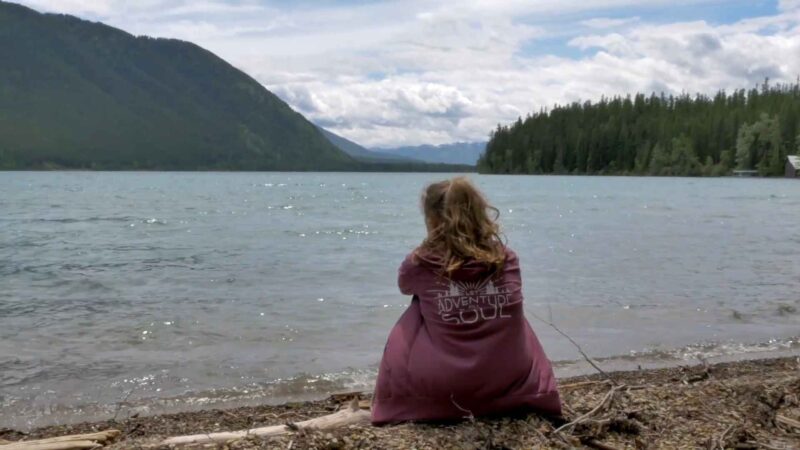 Person with long hair wearing a maroon hoodie that says “Adventure Soul,” sitting by the lake with forested hills and mountains in the background