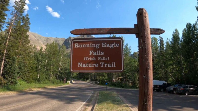 Wooden sign reading "Running Eagle Falls (Trick Falls) Nature Trail" against a backdrop of trees, mountains, and a clear blue sky