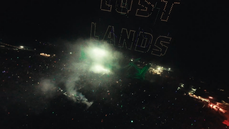 A massive crowd gathers at night under a glowing "Lost Lands" drone display, surrounded by smoke and stage lights