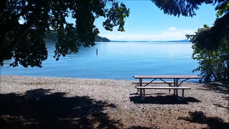 A picnic table sits on a shaded pebble beach overlooking the calm blue water at Yellow Bay State Park