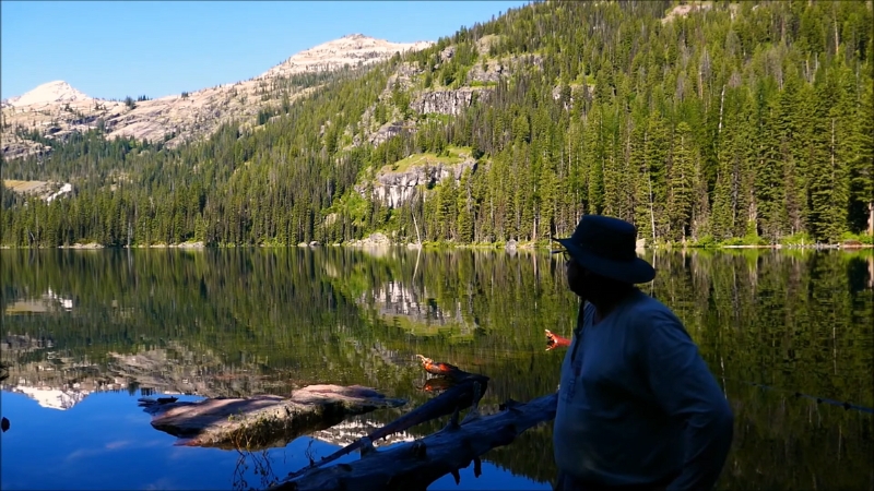 Man stands by Marshall Lake with forest and mountain peaks reflected in the water