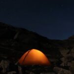 A brightly lit tent stands in the mountains at night, surrounded by dark silhouettes of peaks under a starry sky