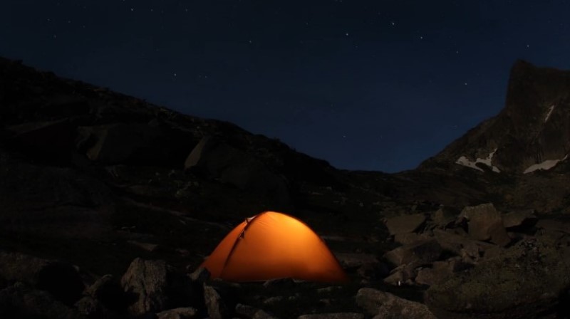 A brightly lit tent stands in the mountains at night, surrounded by dark silhouettes of peaks under a starry sky