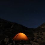 A brightly lit tent stands in the mountains at night, surrounded by dark silhouettes of peaks under a starry sky