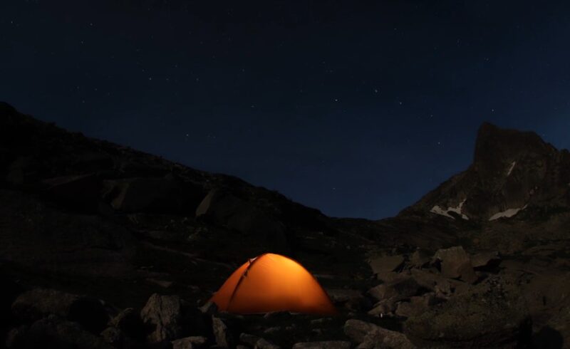A brightly lit tent stands in the mountains at night, surrounded by dark silhouettes of peaks under a starry sky