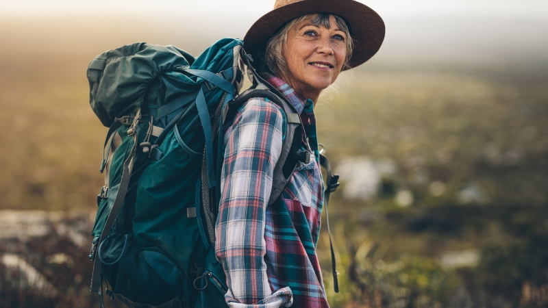 A smiling hiker with a backpack and hat stands on a scenic trail