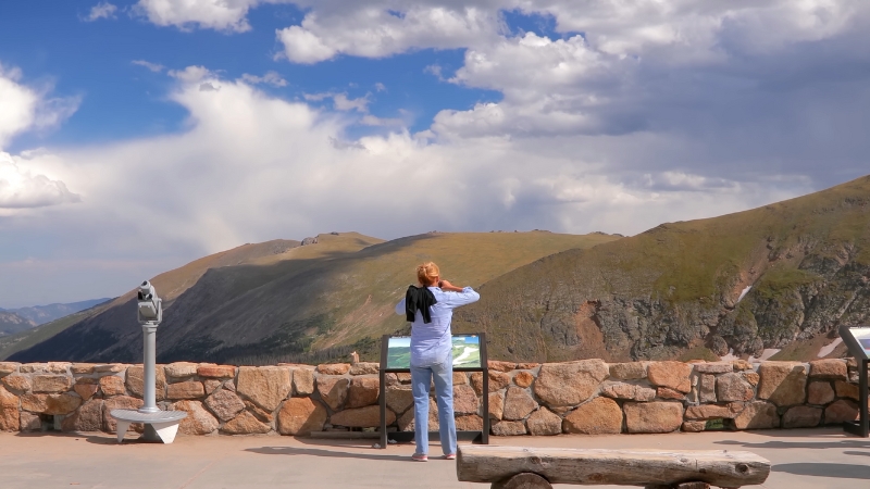 A visitor looks out over the Rocky Mountains from a scenic viewpoint with information boards and a telescope