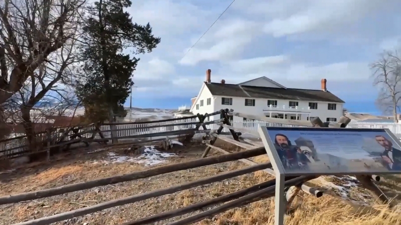 A snowy scene near a historic building in Deer Lodge with a wooden fence and an informational sign in the foreground