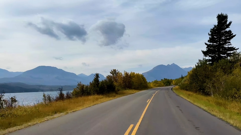 A two-lane road runs beside a lake and forested mountains in Glacier National Park, Montana