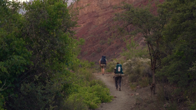 Two backpackers walking on a narrow trail through dense greenery toward red canyon walls