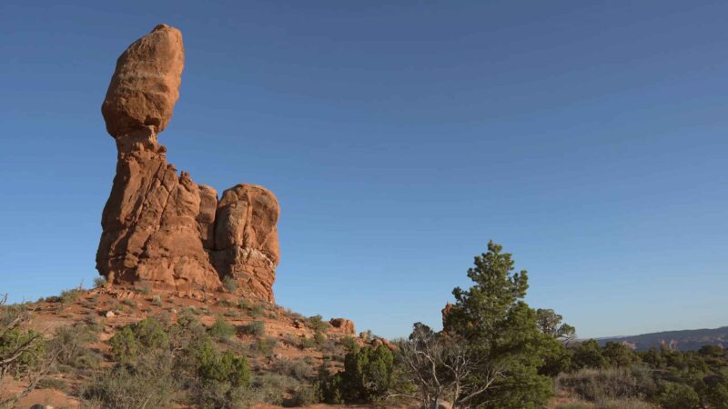 Tall balanced red rock formation rising above desert shrubs under a clear blue sky