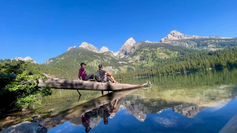 Two hikers sitting on a fallen log over a clear mountain lake with Teton peaks reflected in the water