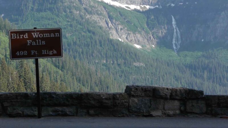 A roadside sign reading “Bird Woman Falls – 492 Ft. High” with a tall, narrow waterfall cascading down a forested mountainside in the distance