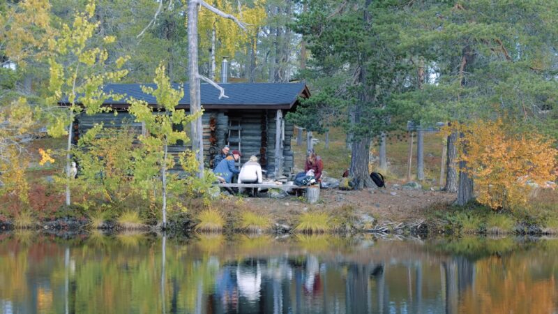 A group rests beside a log cabin near a calm forest lake