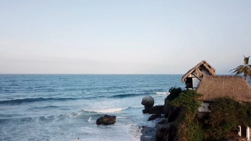 Ocean waves beside rustic thatched-roof cottages on a rocky tropical coast